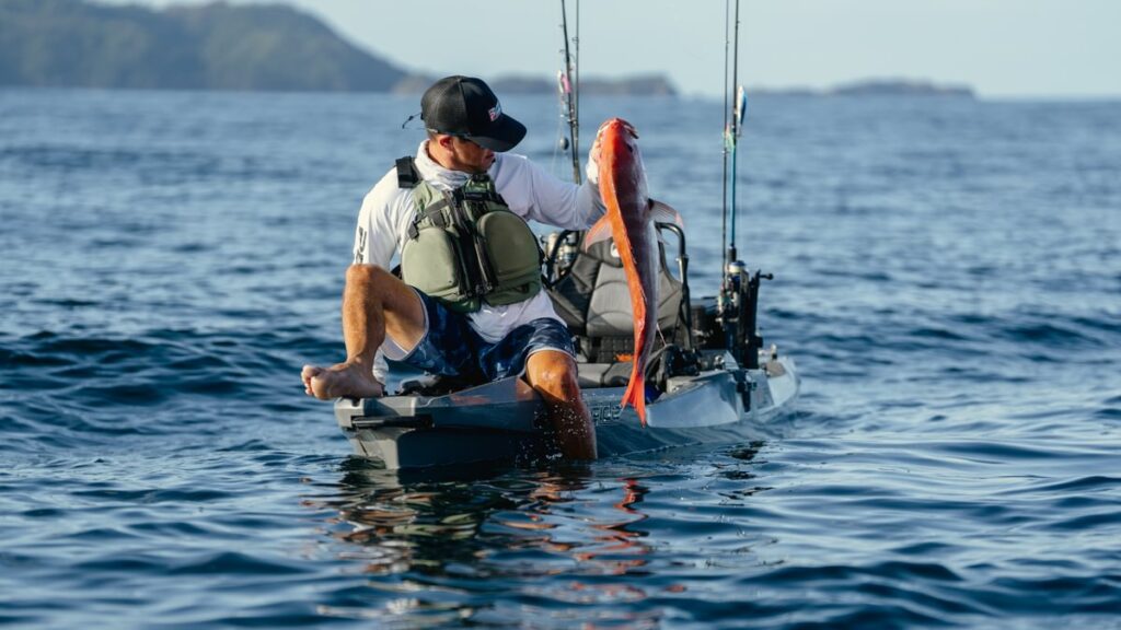Wędkarz bezpiecznie wyciąga rybę z wody, siedząc na stabilnym kajaku wędkarskim Bonafide PWR 129. Pokaz stabilności markowego kadłuba podczas finałowej fazy holu