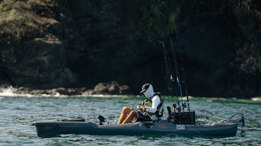 Wędkarz bezpiecznie wyciąga rybę z wody, siedząc na stabilnym kajaku wędkarskim Bonafide PWR 129. Pokaz stabilności markowego kadłuba podczas finałowej fazy holu