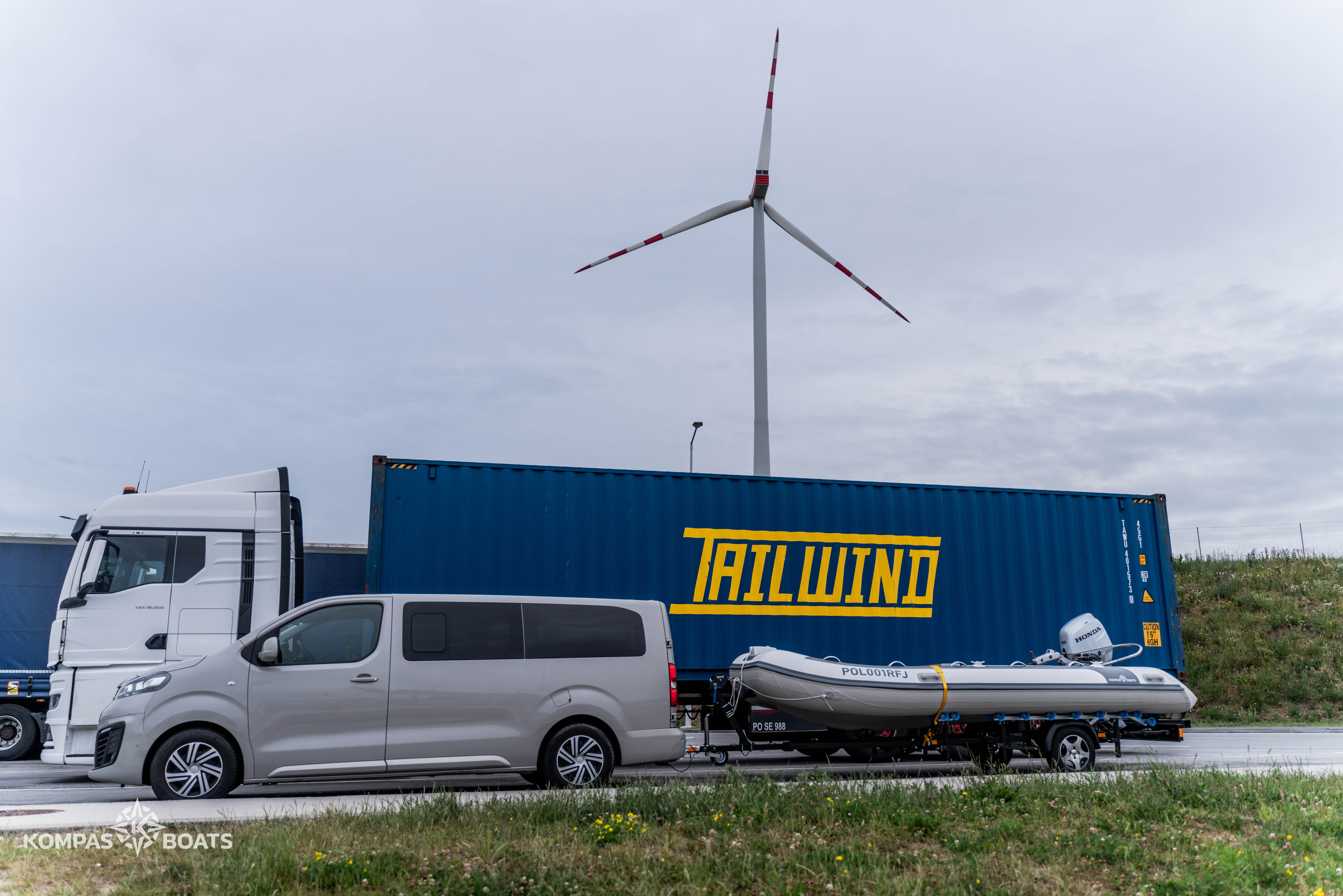 Duży ponton marki Kompas Boats na przyczepie podłodziowej, holowany przez samochód typu van, stojący obok ciężarówki w celu porównania gabarytów.