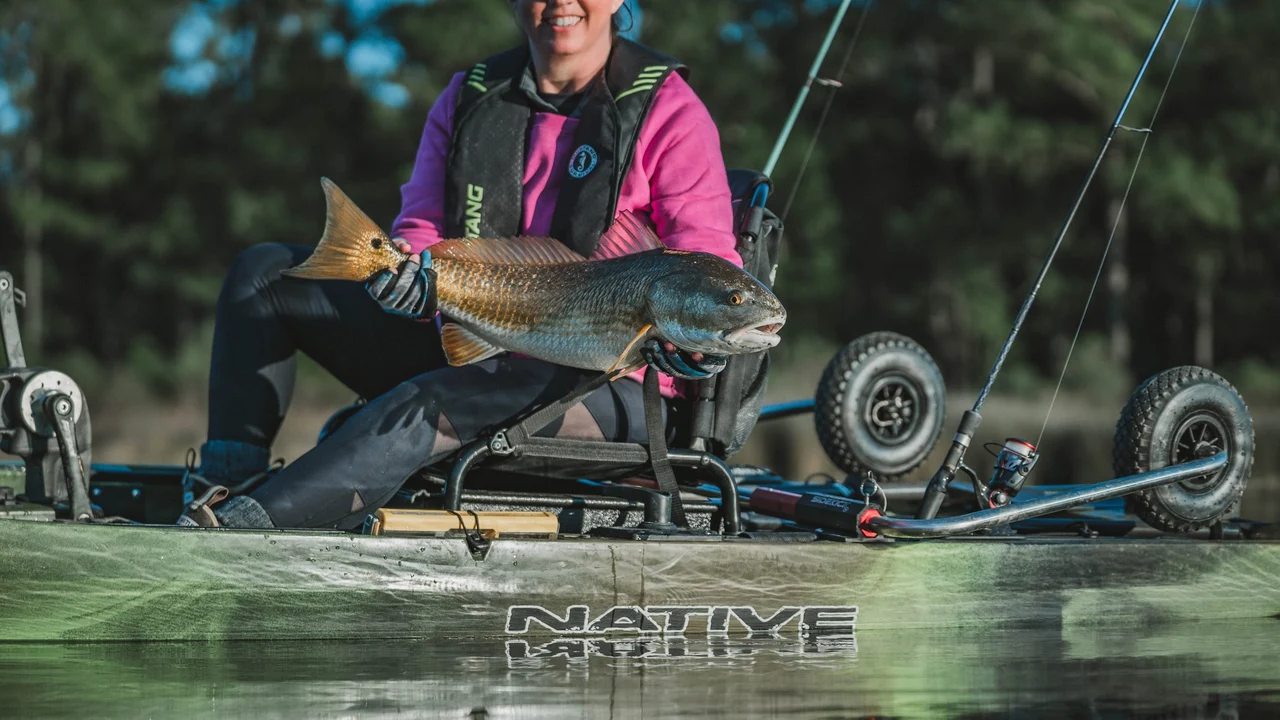 Wędkarka na kajaku Native Watercraft prezentująca złowioną rybę z widocznym na rufie podniesionym wózkiem SideKick Wheel Transport System.