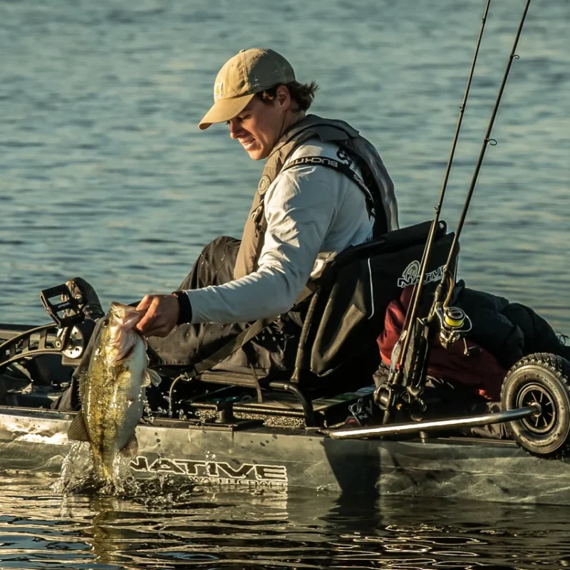 Wędkarz na kajaku Native Watercraft trzymający rybę z widocznym podniesionym kołem zintegrowanego wózka SideKick Wheel Transport System