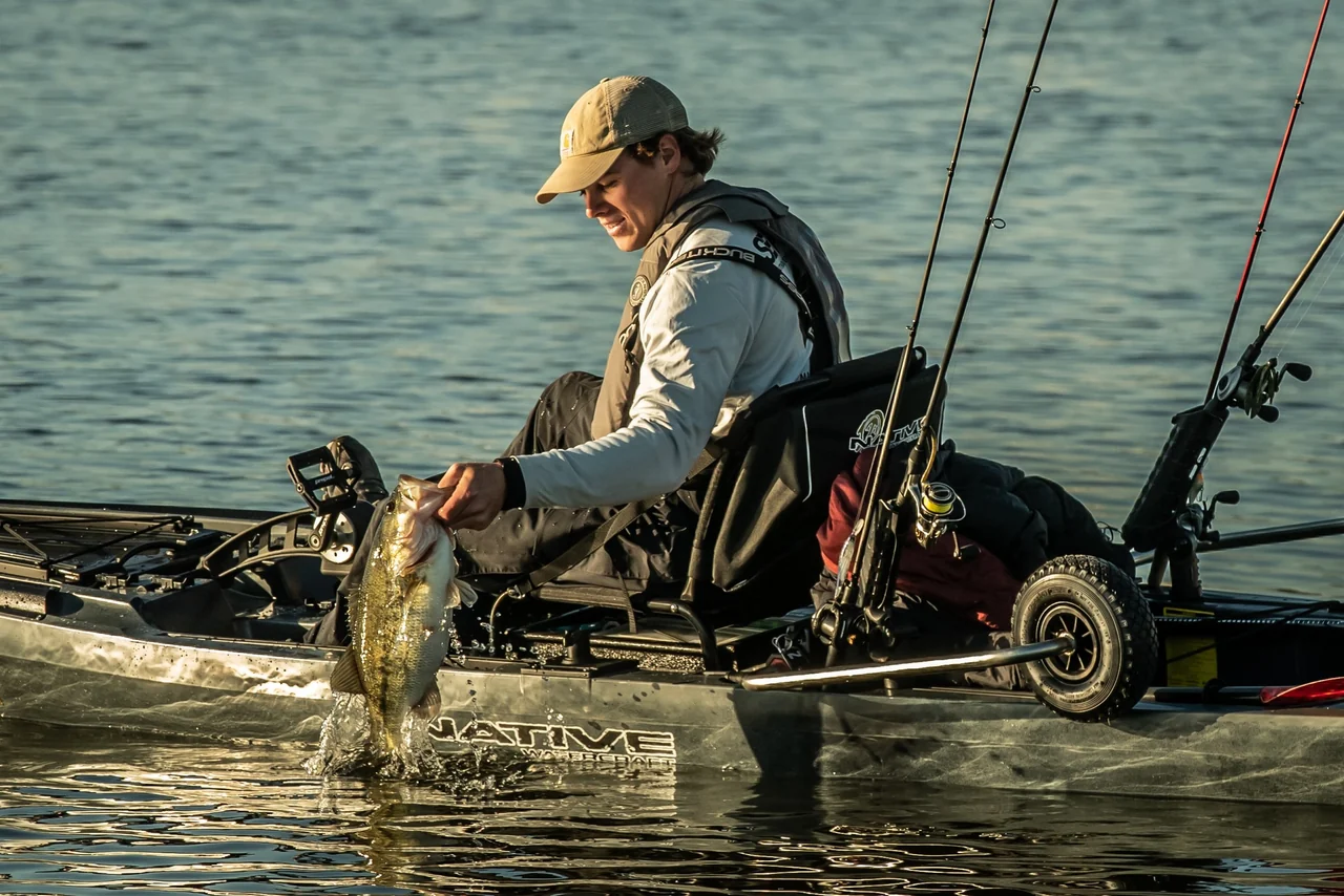 Wędkarz na kajaku Native Watercraft trzymający rybę z widocznym podniesionym kołem zintegrowanego wózka SideKick Wheel Transport System