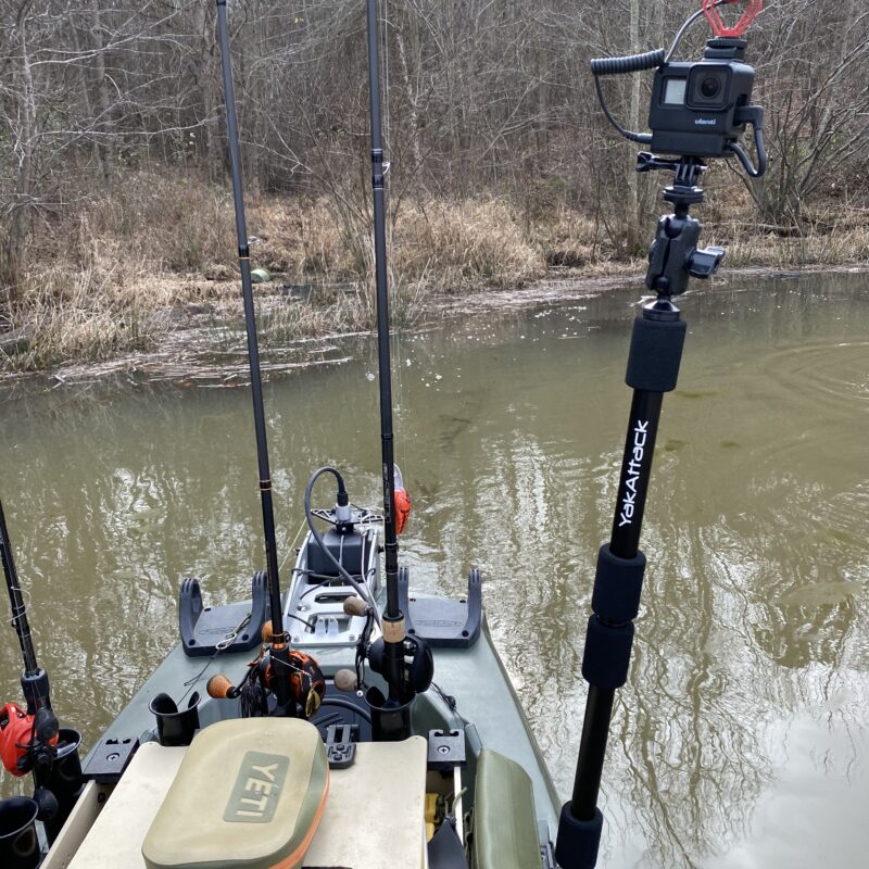 Uchwyt do kamery na kajak YakAttack PanFish Pro™ Camera Mount z zamontowaną kamerą GoPro na rufie uzbrojonego kajaka wędkarskiego na tle wody i lasu