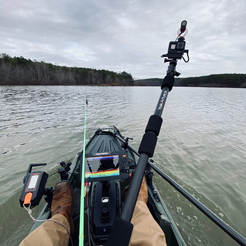 Widok z pokładu kajaka wędkarskiego na trzymany w dłoni uchwyt do kamery YakAttack PanFish Pro™ Camera Mount z kamerą GoPro i echosondą Raymarine na tle jeziora